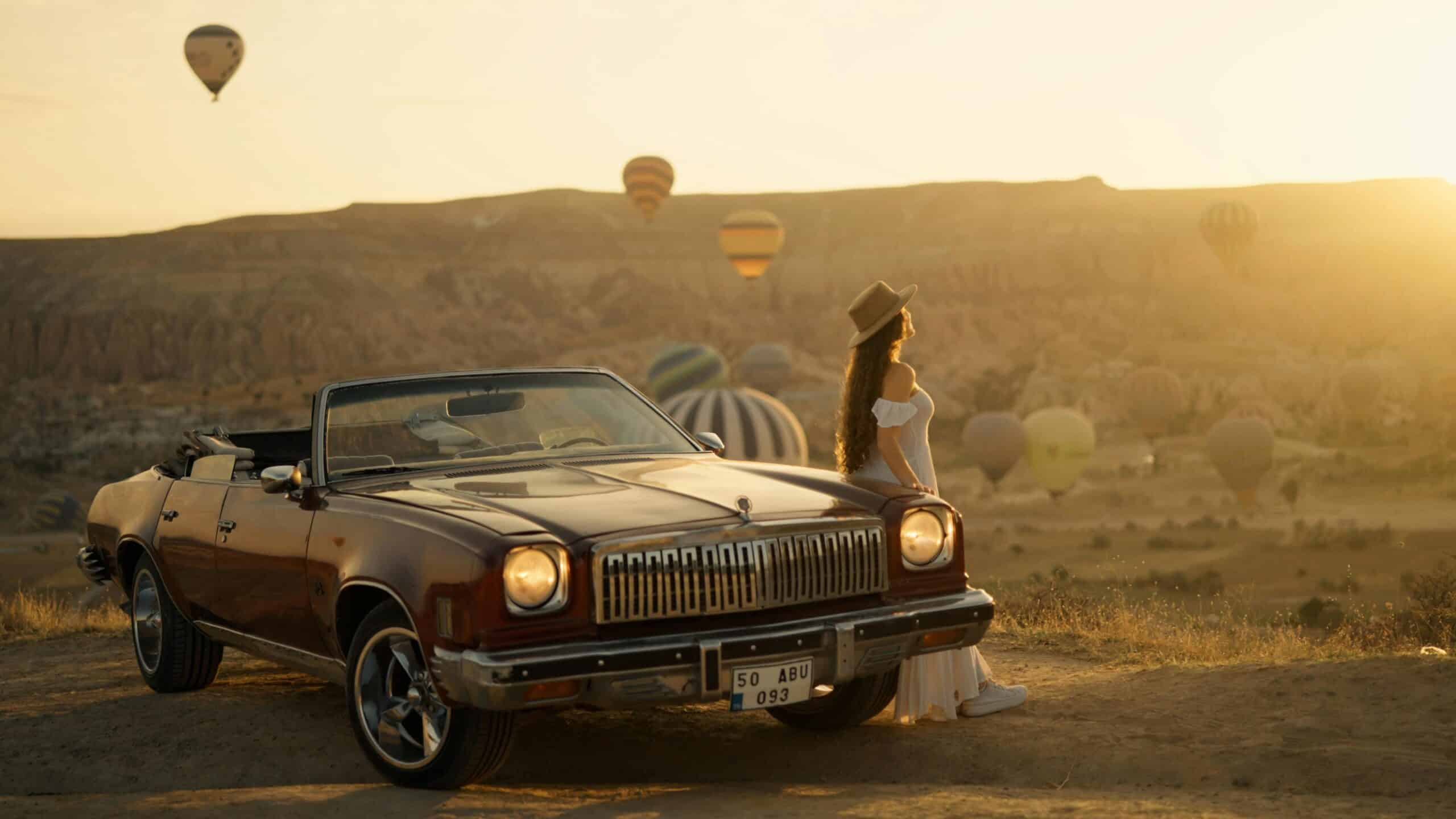 Woman standing beside a vintage car at sunrise with hot air balloons over Cappadocia, Turkey